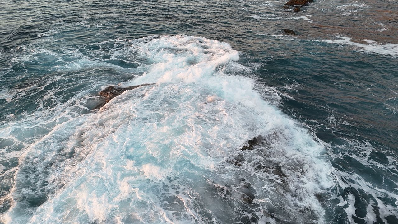 Aerial view of surf breaking over dark coastal rocks.
